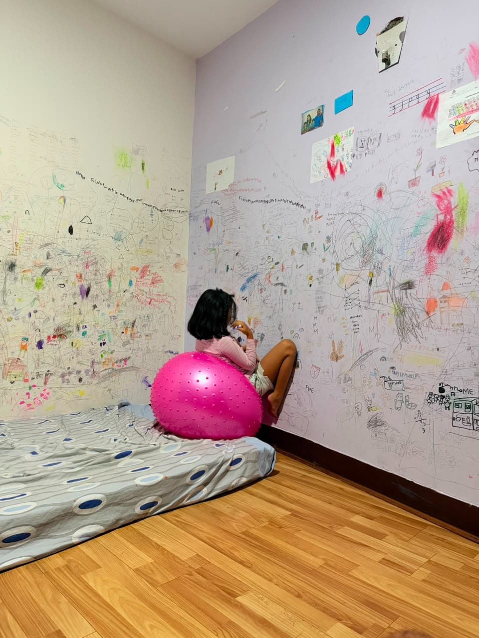 A girl sitting on a pink exercise ball and making art on walls filled with her drawings.
