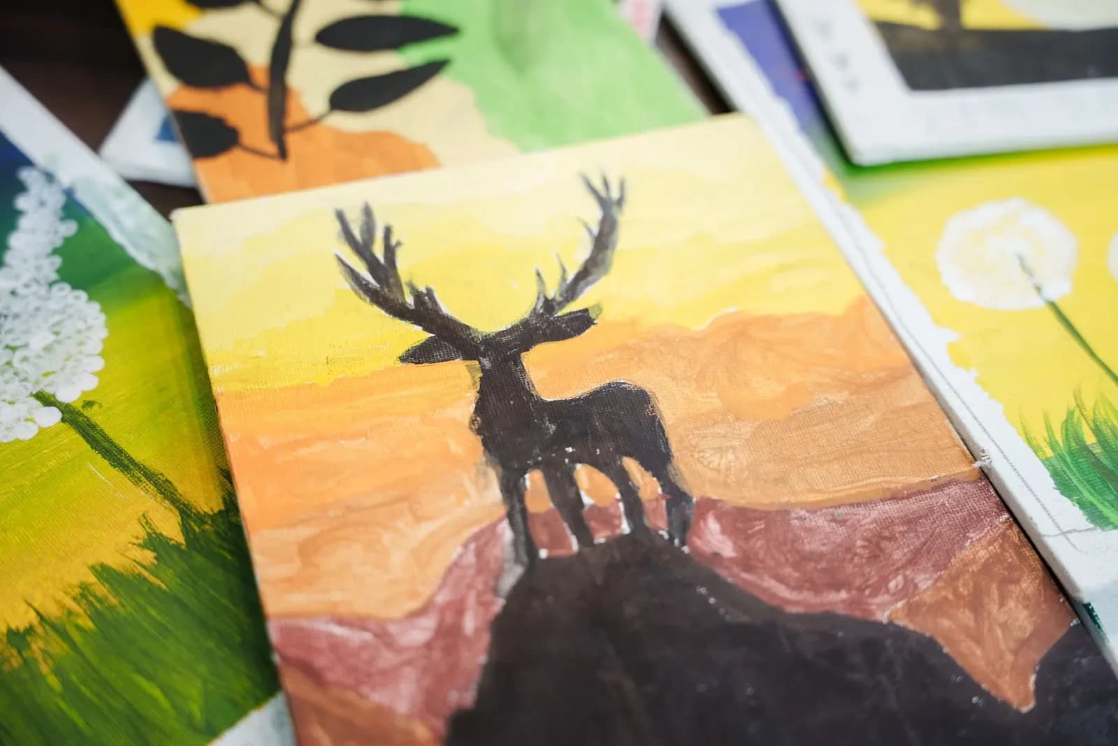 A painting of a dark deer silhouette with antlers standing on a rocky ledge against an orange and yellow sunset sky.