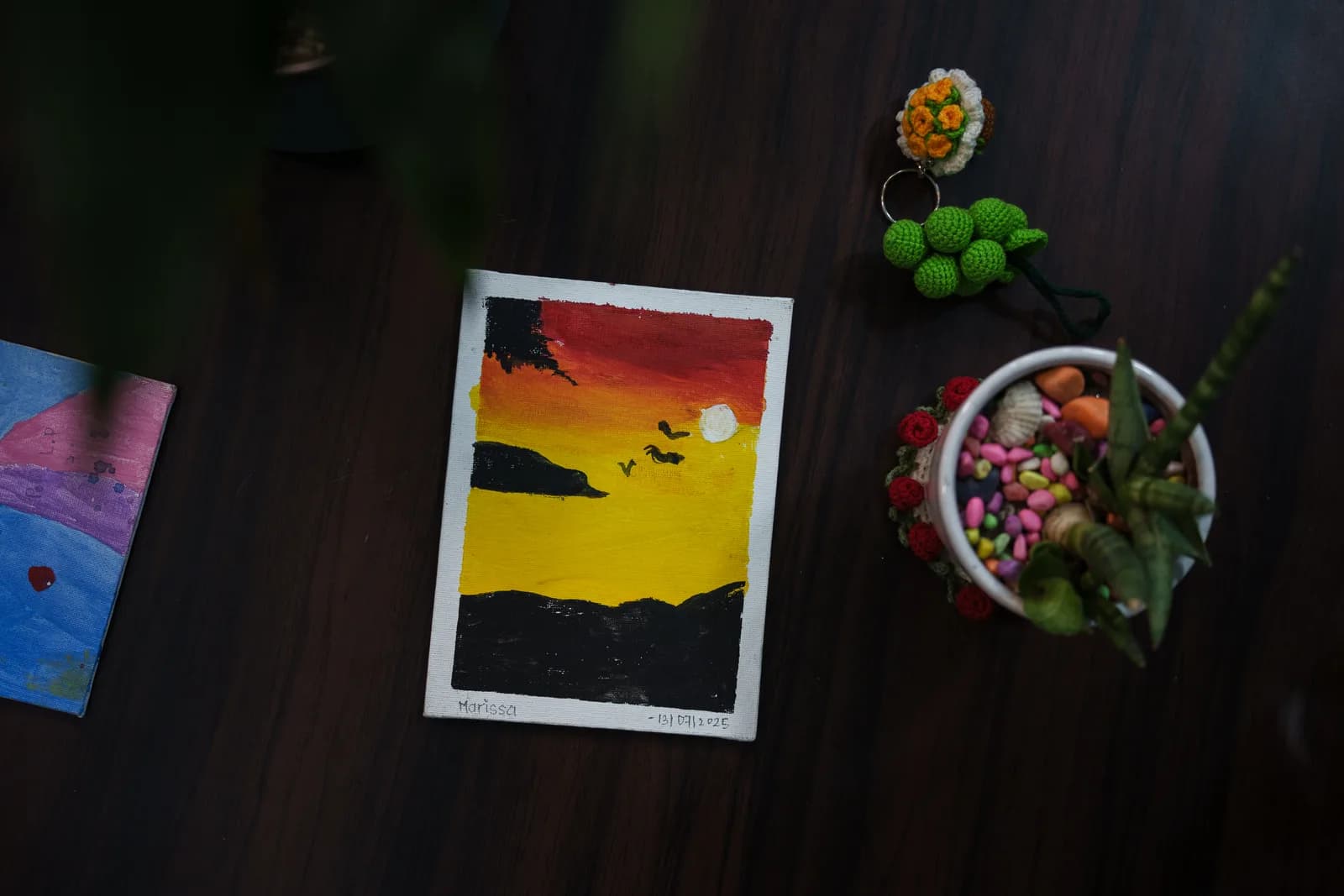 A yellow sunset painting photographed beside small flowers and colourful ornaments.