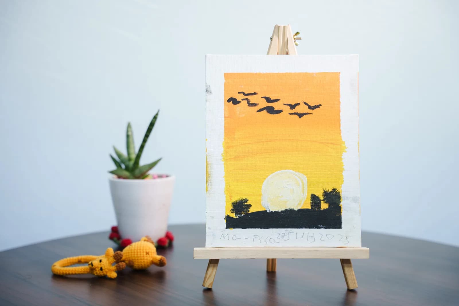 An orange sunset and flying bird painting on an easel beside a green plant.