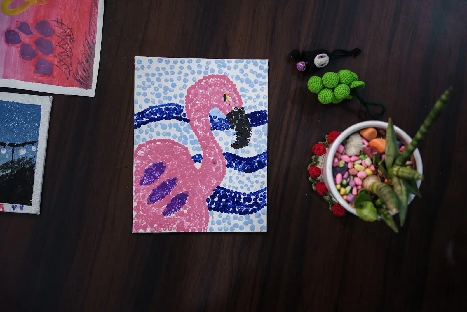 A dotted painting of a pink flamingo in front of blue and white wave stripes, laid on a dark table beside other paintings.